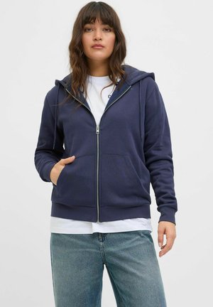 Navy zip-up hoodie with a drawstring hood, front pockets, and ribbed cuffs, worn over a white t-shirt and blue jeans. Smooth fabric texture.