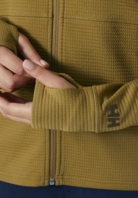 Chaqueta verde oliva con un patrón texturizado, que presenta una cremallera y orificios para los pulgares en los puños, con un logotipo en contraste impreso en el brazo.