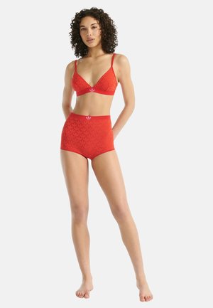 Mujer de pie descalza, vistiendo un sujetador corto rojo con estampado y braguitas de talle alto con un pequeño logo en la cintura, fondo claro.