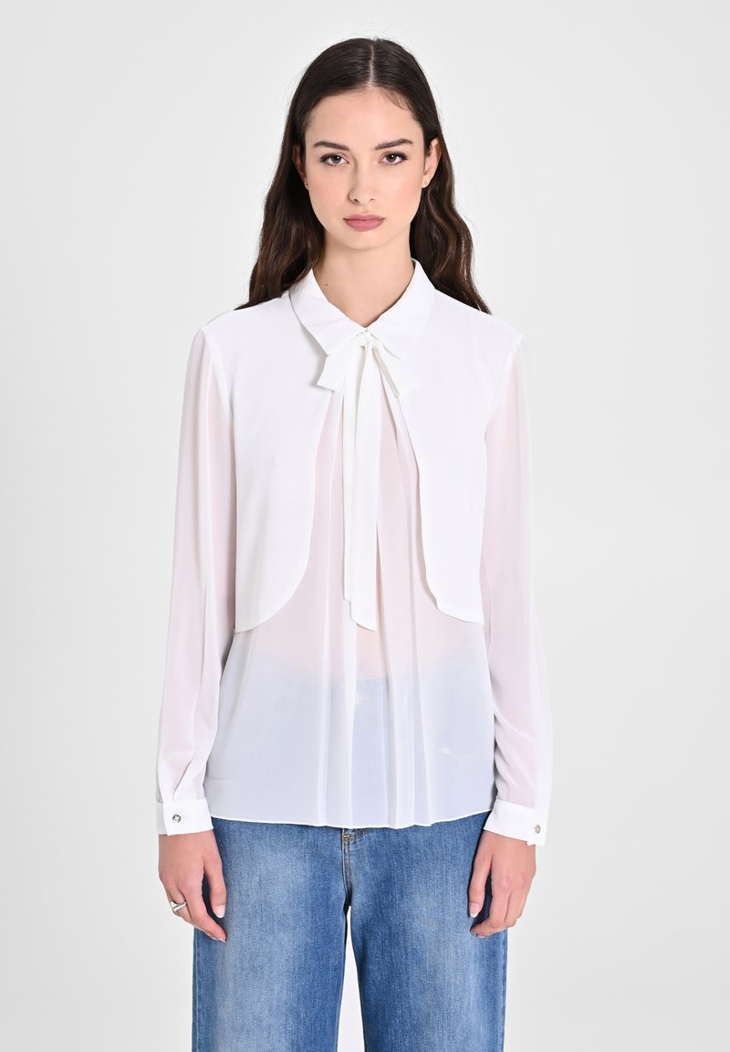 Blusa bianca leggera con colletto e dettaglio del fiocco. Caratterizzata da maniche lunghe e un design frontale a strati, indossata sopra jeans in denim.