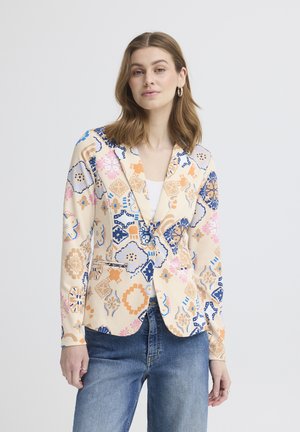 Kvinde iført en cremefarvet blazer med flerfarvede geometriske blomsterprint og blå jeans, stående foran en lys baggrund.