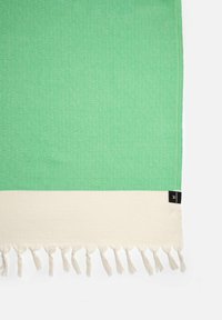 Tessuto verde a motivo con un design a diamante, caratterizzato da un bordo color crema e nappine decorative lungo il bordo.