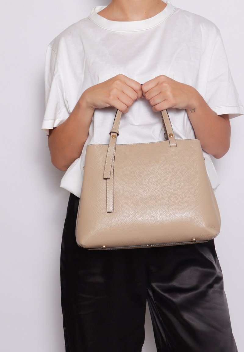 Borsa a mano beige con texture, manici corti, dettagli lisci e base arrotondata. Esposta su sfondo bianco, tenuta da una persona.