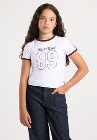 Ragazza con lunghi capelli ricci che indossa una maglietta bianca con la scritta "New York 89" e pantaloni scuri a righe, in piedi con una mano in tasca.