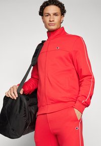 Chándal rojo con cremallera y detalles en blanco, fabricado en un tejido suave. El modelo posa con un bolso de viaje negro, mostrando el diseño deportivo del atuendo.