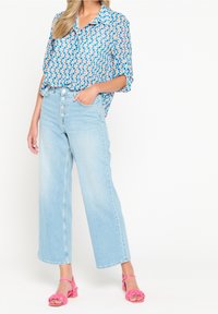 Blusa blu a maniche corte con motivo circolare, abbinata a jeans a gamba larga blu chiaro. Il modello indossa sandali rosa con cinturino.