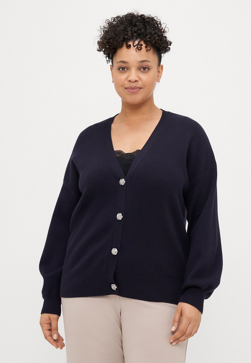 Mulher com cabelo encaracolado, vestindo um cardigã azul escuro com botões decorativos sobre uma blusa de renda preta e calças beiges, de pé contra um fundo branco.