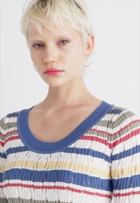 Črtasta pletena jopica z modrim ovratnikom. Ima zelene, rdeče, rumene in bele črte. Material je teksturiran in rahlo prosojen.