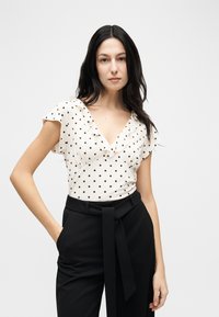 Γυναίκα με μακριά σκοτεινά μαλλιά που φοράει λευκό πουκάμισο με πουα και βολάν στο λαιμό και ψηλόμεσα μαύρα παντελόνια με δέσιμο στη μέση.