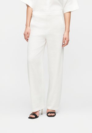 Kvinde iført højtaljede løse hvide bukser kombineret med sorte sandaler med åben tå og tynd gylden stiletthæl.