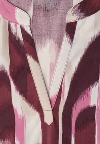 Détail du col d'une chemise en tissu à motifs rose, grenat et crème, avec une ouverture en V et des bords cousus.