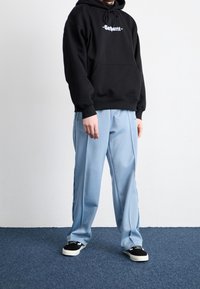 Felpa nera con cappuccio, tasca frontale e logo, abbinata a pantaloni a pieghe azzurri chiari. Le calzature presentano una combinazione di colori nero e bianco.