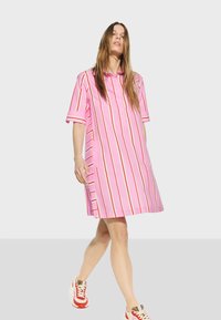 Růžové pruhované šaty s krátkým rukávem, zapínání na knoflíky vpředu a boční kapsy. Pruhy mají červené a bílé akcenty. V kombinaci s červenými teniskami.