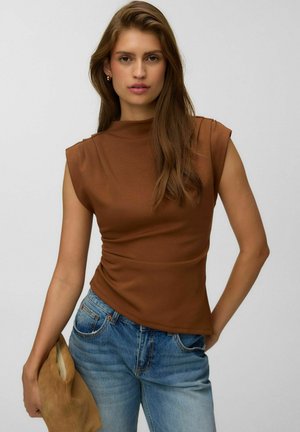 Donna con lunghi capelli castani che indossa un top marrone senza maniche e jeans blu, che tiene in mano una pochette in suede color cammello su uno sfondo semplice.