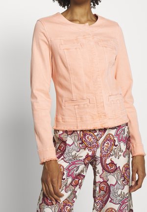 Vrouw draagt een lichtroze spijkerjack met rafelige randen en een witte broek met paarse en oranje paisleypatronen.