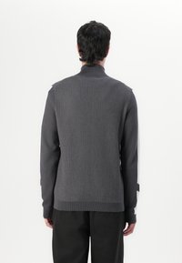 Grau gestrickter Pullover mit hohem Kragen, ausgestattet mit strukturiertem Rippenmuster und dezenten Mustern im Schulterbereich. Kombiniert mit dunklen Hosen.