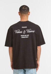 Sort bomulds T-shirt med stort hvidt tekst p�å ryggen: "PEAGADOR," "Værdier & Visioner," og "Disciplin og Konsistens." Korte ærmer.