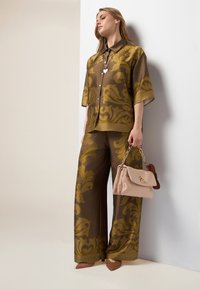 Braunes Satinhemd und weite Hose mit gelben Blumenmustern. Beiger Handtasche mit goldenen Beschlägen und braunen Akzenten. Braune geflochtene Absätze.