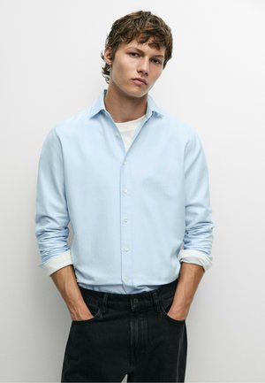 Giovane uomo con capelli castani indossa una camicia azzurro chiaro abbottonata sopra una maglietta bianca e jeans neri, in piedi con le mani nelle tasche davanti a un muro bianco.