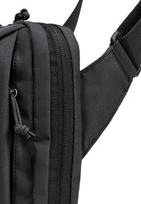 Gros plan sur la sangle d'un sac à dos en tissu noir avec une fermeture éclair et une bretelle d'épaule rembourrée ajustable sur un fond blanc.