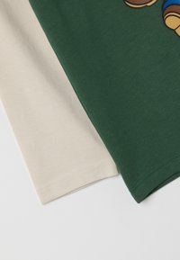 Twee T-shirt mouwen die elkaar overlappen: één donkergroen, de ander lichtbeige. Beide zijn gemaakt van glad katoen, met zichtbare stiksels aan de randen.