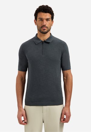 Man met krullend haar, gekleed in een donkergrijs korte mouwen gebreid poloshirt en lichtbeige broek, staand tegen een witte achtergrond.