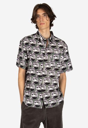 Junger Mann trägt ein kurzärmliges Hemd mit Knöpfen und einem sich wiederholenden abstrakten schwarz-weißen Gesichts-Muster, kombiniert mit dunklen Hosen.