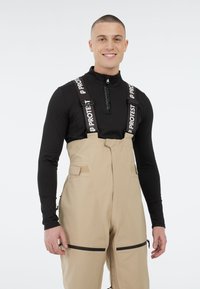Beige skibroeken met verstelbare banden, ritszakken en een slim-fit ontwerp, gecombineerd met een aansluitend zwart longsleeve shirt.