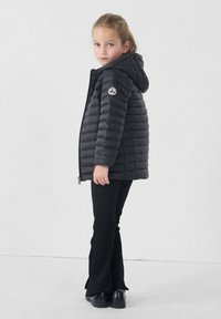 Giacca a piumino nera con cappuccio e toppa con logo, presenta una chiusura frontale con cerniera e una texture trapuntata orizzontale. Abbinata a pantaloni neri svasati.