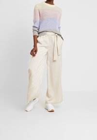 Lichtbeige wijde broek met een gestrikt taille-detail, gecombineerd met een pastelkleurige gestreepte gebreide trui in tinten paars, roze en geel.