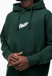 Sweatshirt à capuche vert avec une coupe décontractée, présentant un logo brodé blanc sur la poitrine et des poignets en côtes. Texture de tissu lisse.