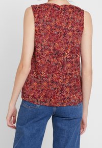 Bagview af en ærmeløs top i rød med et blomsterprint, der har orange, gul og sort accent, parret med høj detaljerede blå jeans.