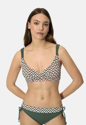 Joven mujer con bikini verde y estampado con diseño de zigzag y tirantes trenzados, de pie con las manos detrás de la espalda.