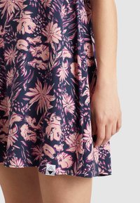 Mekko, jossa on merivoimien värinen kukkakuosi, jossa on suuria vaaleanpunaisia ja valkoisia kukkia. Kangas on pehmeä ja siinä on virtaava muotoilu. Mukana on pieni brändilogo.