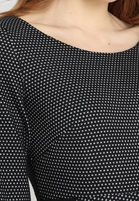 Top en tricot noir et blanc à motifs, avec un large décolleté et un tissu texturé, affichant de petits carrés dans tout le design.