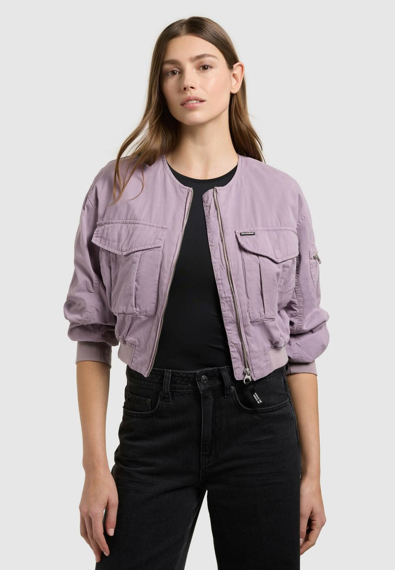 Jeune femme aux cheveux bruns raides portant une veste bomber courte lavande par-dessus un t-shirt noir et un jean noir, debout devant un fond gris.