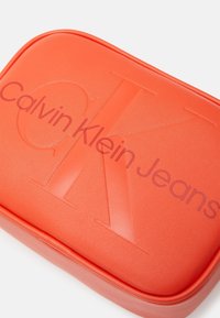 Pochette arancione Calvin Klein Jeans con superficie liscia e testurizzata, caratterizzata da un logo "CK" in rilievo e dal nome del marchio in rosso scuro.