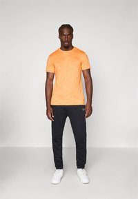 Πορτοκαλί κοντομάνικη μπλούζα με διακριτικό υφασμάτινο μοτίβο, συνδυασμένη με μαύρο παντελόνι και λευκά αθλητικά παπούτσια. Απλός σχεδιασμός, άνετη εφαρμογή.