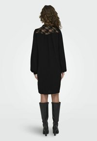 Czarna sukienka z koronkowym panelem z tyłu, długimi rękawami i luźnym krojem. Połączona z czarnymi skórzanymi kozakami sięgającymi za kolano. Gładka tekstura.