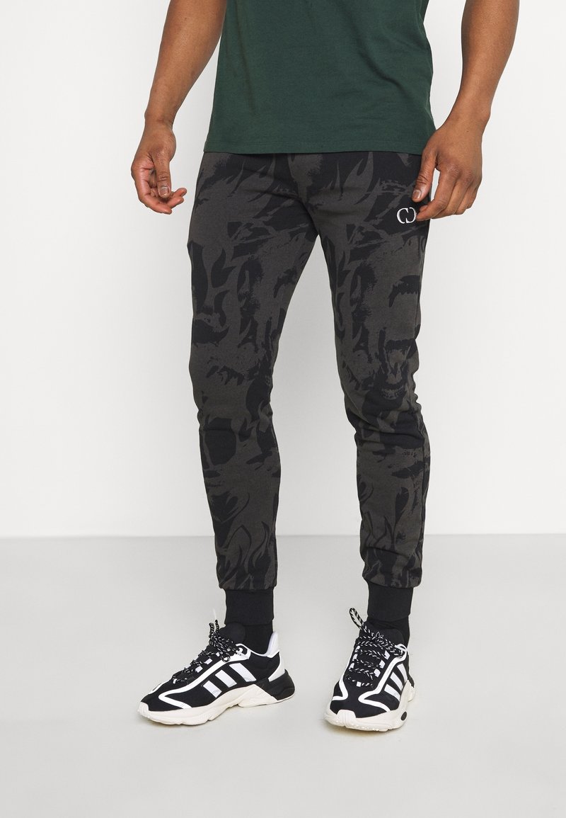 Zwarte joggers met een camouflagepatroon, aansluitende enkelmanchetten en een geborduurd logo aan de zijkant. Gdragend met zwart-witte sneakers.