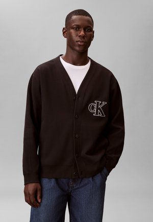 Jeune homme portant un cardigan noir boutonné avec le logo "cK" blanc, un t-shirt blanc et un jean bleu, debout devant un fond gris uni.