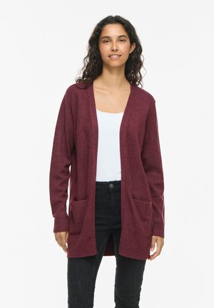 Kvinde med krøllet hår iført en lang bordeaux cardigan over en hvid top og sorte bukser, stående mod en ensfarvet hvid baggrund.