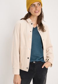 Beige corduroy jack met knoopsluiting en zichtbare stiksels, gedragen over een teal shirt. Zwarte broek maakt de outfit compleet. Gebreide mosterdkleurige beanie.