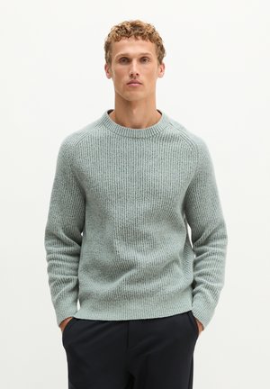 Mann mit lockigem Haar, der einen hellgrauen gerippten Strickpullover und dunkle Hose trägt, steht mit den Händen in den Taschen vor einem schlichten Hintergrund.