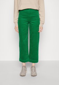 Pantalons larges en velours côtelé vert avec des nervures verticales, taille haute et ourlets, associés à des chaussures de sport beiges.