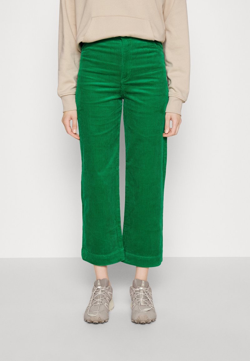 Pantalons larges en velours côtelé vert avec des nervures verticales, taille haute et ourlets, associés à des chaussures de sport beiges.