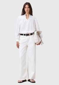 Camicia bianca a maniche lunghe con chiusura a bottoni, dettagli plissettati, abbinata a jeans bianchi a gamba larga e cintura nera. Tiene in mano una borsa chiara.