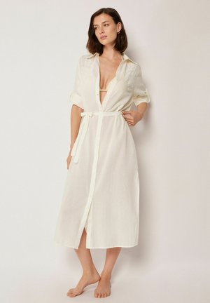 Frau in einem weißen, langen, geknöpften Kleid mit hochgekrempelten Ärmeln und gebundener Taille, die barfuß vor einem schlichten hellen Hintergrund steht.