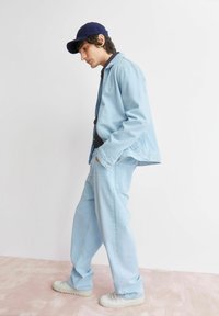 Hellblaue Jeansjacke und Hose, kombiniert mit weißen Sneakers und einer marineblauen Kappe. Das Outfit hat eine lässige Passform und klassische Designdetails.