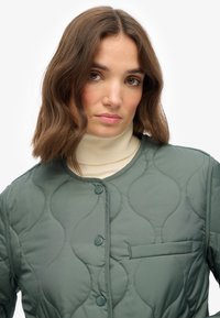 Gequiltede groene jas met ronde stikpatronen, voorste knopen en een klein borstzakje. Gedragen over een crèmekleurige coltrui.